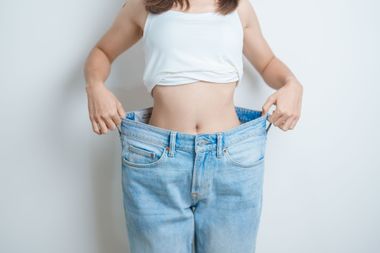 Woman holding oversized blue jeans around her waist against a plain background